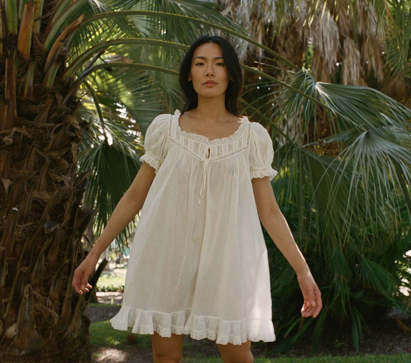 Female model standing in garden wearing white cotton DÔEN dress with short puffed sleeves trimmed with lace and ruffles